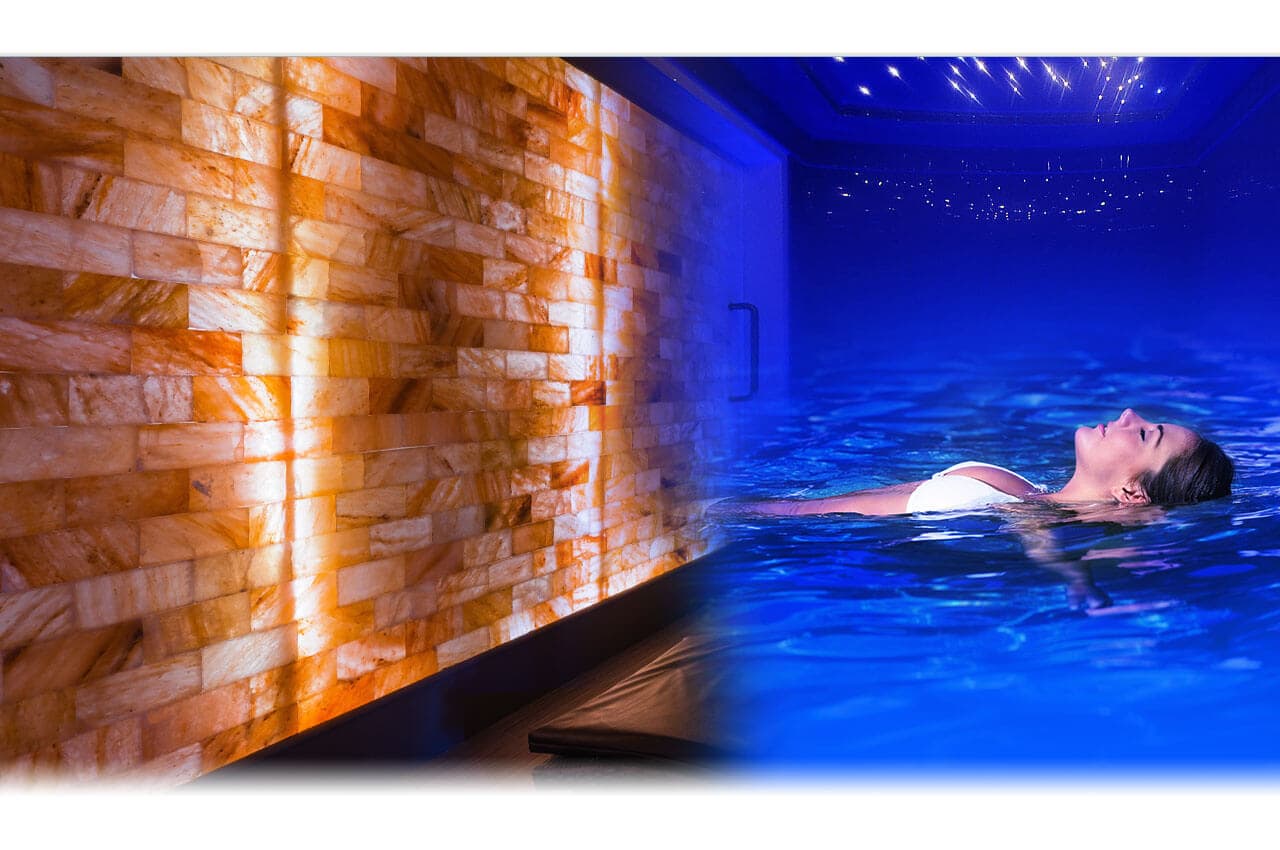 Person floating in illuminated blue salt water tank with warm orange brick wall beside