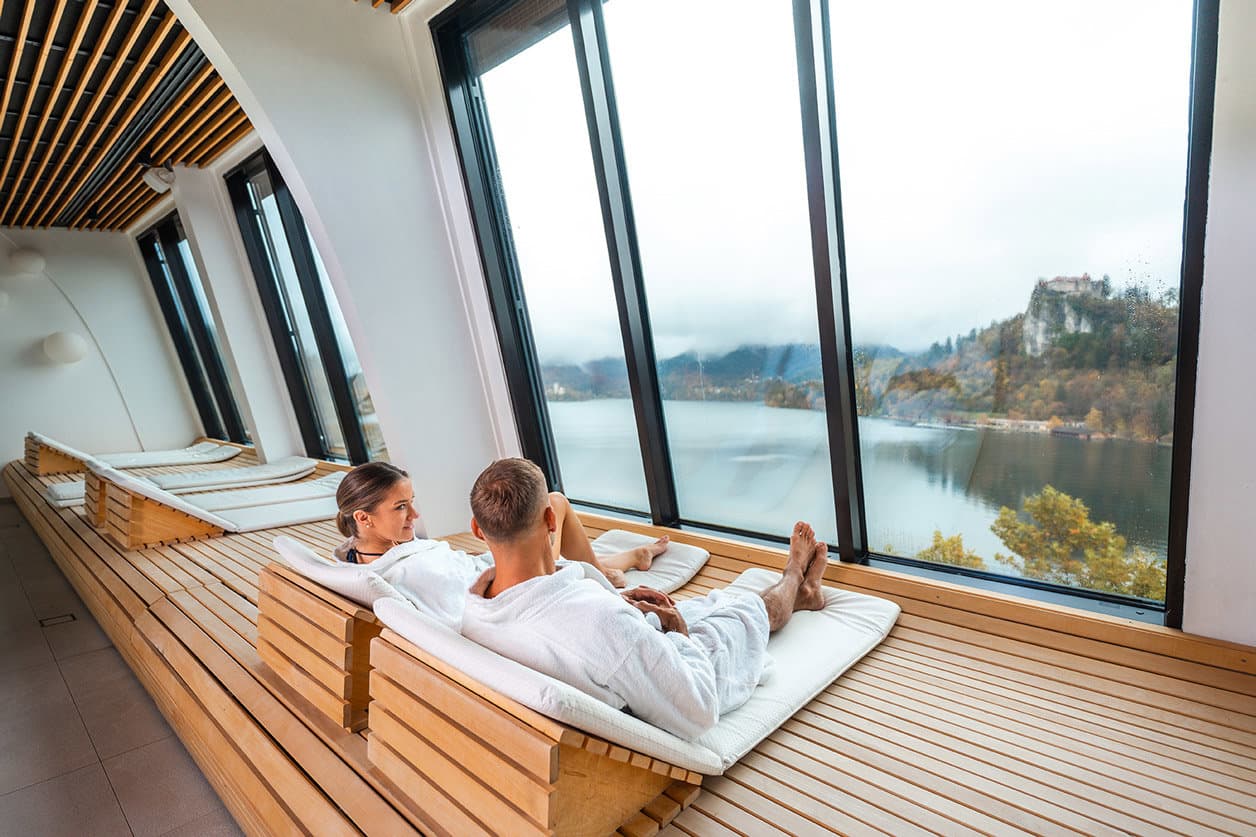 Couple relaxing on wooden loungers overlooking a lake with mountains in a spa room with large windows