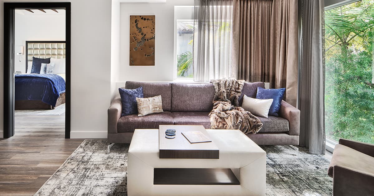 Modern suite living area with gray sofa, white coffee table, and bedroom visible through doorway