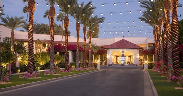 Illuminated driveway lined with palm trees leading to main building at dusk